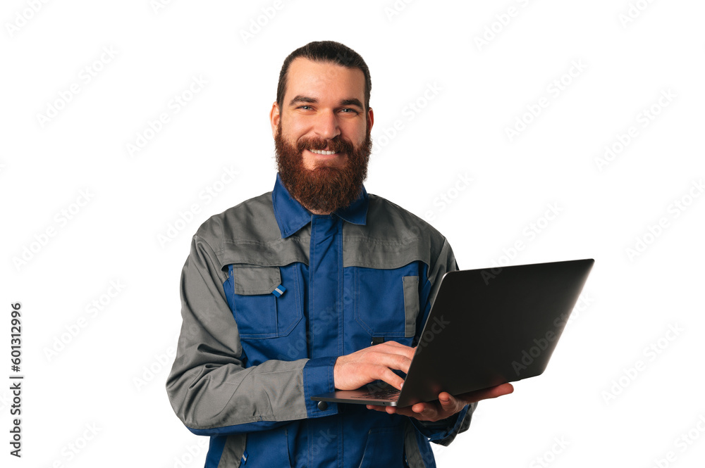 Wide smiling bearded man wearing engineer uniform is holding a laptop over white background. Wide smiling bearded man wearing engineer uniform is holding a laptop over white background.