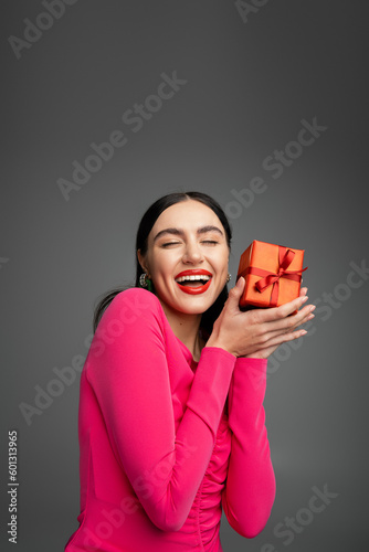 excited and young woman with trendy earrings and brunette hair smiling standing with opened mouth and holding red wrapped present for holiday on grey background