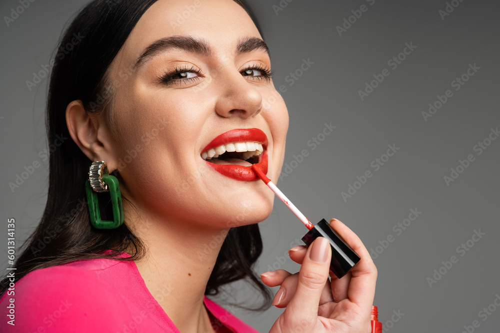 Obraz premium excited woman with shiny brunette hair and trendy earrings applying red lip gloss and smiling while posing in magenta dress isolated on grey background
