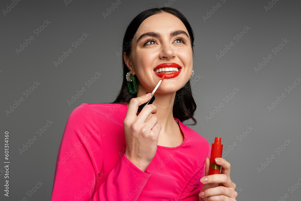alluring young woman with shiny brunette hair and trendy earrings posing in magenta dress while applying red lip gloss before party isolated on grey background alluring young woman with shiny brunette hair and trendy earrings posing in magenta dress while applying red lip gloss before party isolated on grey background