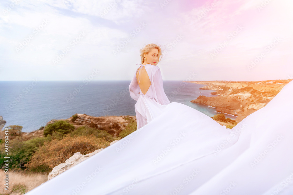 Blonde with long hair on a sunny seashore in a white flowing dress, rear view, silk fabric waving in the wind. Against the backdrop of the blue sky and mountains on the seashore. Blonde with long hair on a sunny seashore in a white flowing dress, rear view, silk fabric waving in the wind. Against the backdrop of the blue sky and mountains on the seashore.