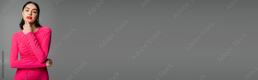 enchanting young woman with shiny brunette hair, trendy earrings, red lips and stylish magenta dress looking at camera and posing on grey background, banner enchanting young woman with shiny brunette hair, trendy earrings, red lips and stylish magenta dress looking at camera and posing on grey background, banner