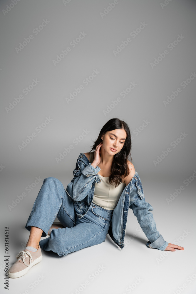 full length of dreamy young woman with long brunette hair and flawless natural makeup posing in stylish blue jeans and denim jacket while sitting on grey background full length of dreamy young woman with long brunette hair and flawless natural makeup posing in stylish blue jeans and denim jacket while sitting on grey background