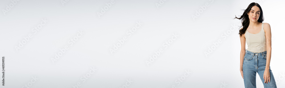 gorgeous woman with brunette hair, perfect skin and natural makeup standing in denim jeans and tank top while looking at camera on white background, banner gorgeous woman with brunette hair, perfect skin and natural makeup standing in denim jeans and tank top while looking at camera on white background, banner