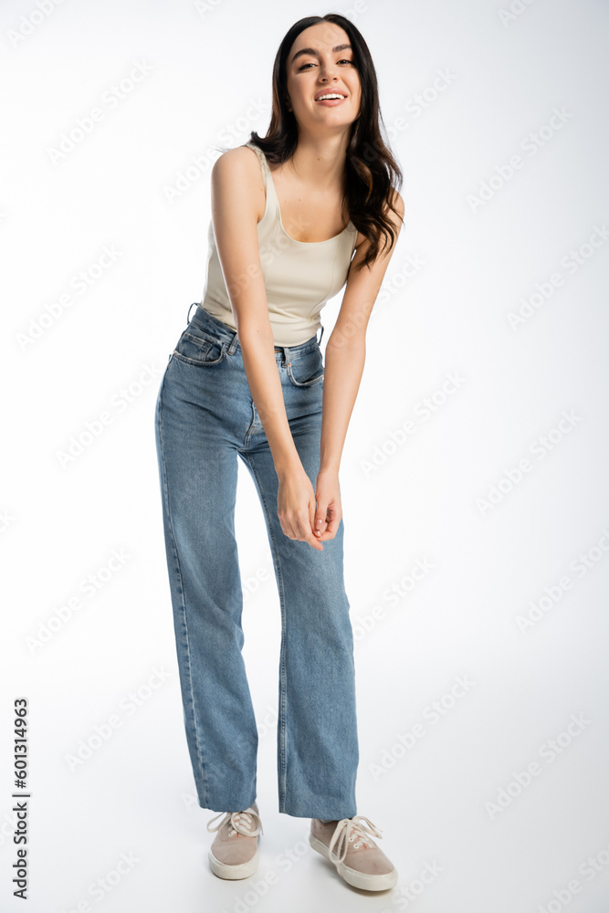 full length of enchanting woman with brunette hair, perfect skin and natural makeup standing in denim jeans and tank top while smiling and looking at camera on white background full length of enchanting woman with brunette hair, perfect skin and natural makeup standing in denim jeans and tank top while smiling and looking at camera on white background