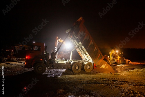Ust-Luga, Leningrad oblast, Russia - November 16, 2021: Road construction site at night and rain. Dump truck unload sand. Excavators flatten ground. Dark sky with orange light.