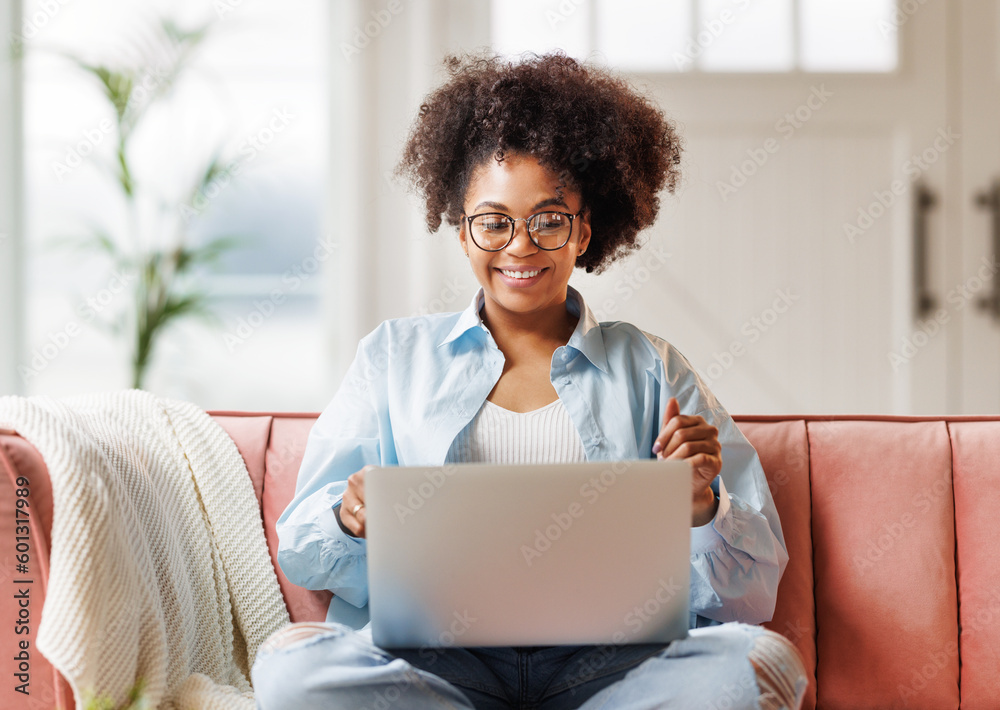 © JenkoAtaman - beautiful young smiling ethnic woman with curly hair uses laptop smiling, gesticulating and talking to online while sitting on sofa © JenkoAtaman - beautiful young smiling ethnic woman with curly hair uses laptop smiling, gesticulating and talking to online while sitting on sofa