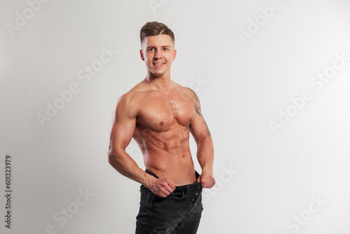 Happy handsome man with smile with muscular body has lost weight and is holding black jeans on a white background. Sports, fitness and weight loss