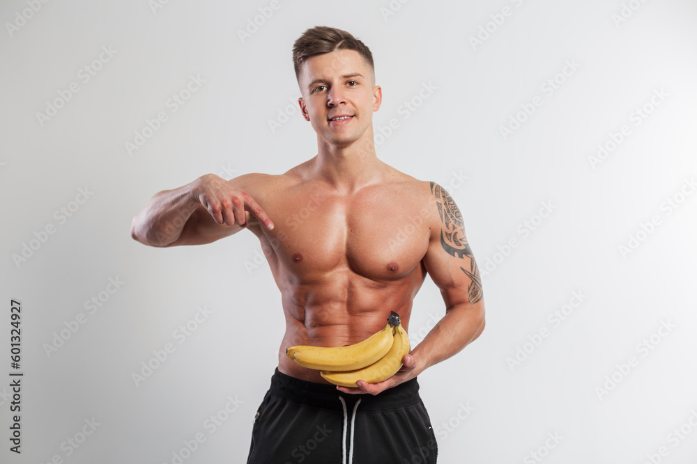 Cool athletic healthy handsome man with muscular body hairstyle holding bananas and pointing with finger in studio. Healthy raw food and vitamins