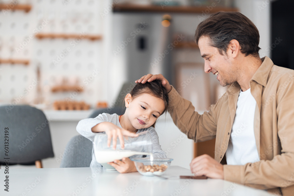 Fototapeta premium A caring father sits with his daughter at the dining table while she makes cereal with milk.