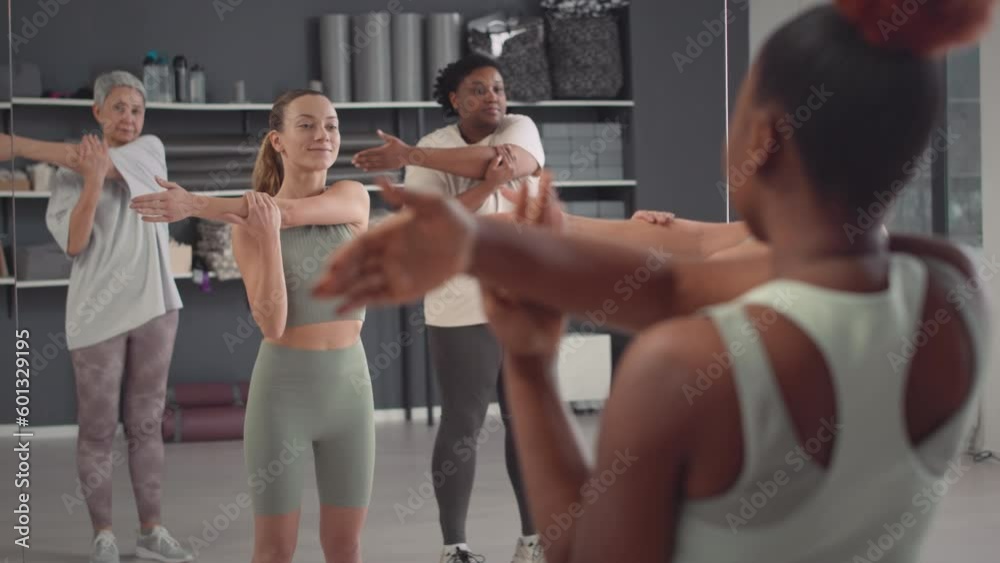 Group of diverse women and their coach stretching their arms while standing in front of full length mirror in spacious bright studio during group fitness class Group of diverse women and their coach stretching their arms while standing in front of full length mirror in spacious bright studio during group fitness class