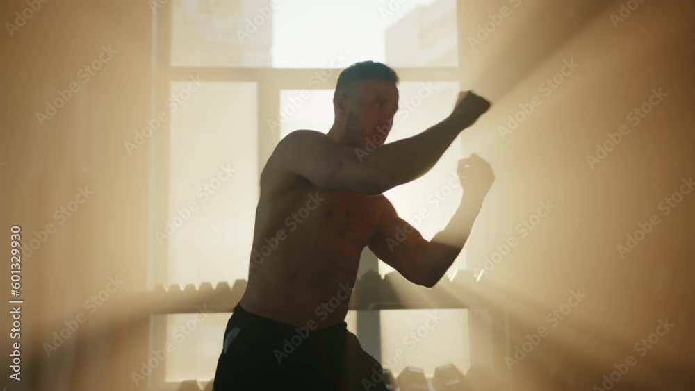 Middle shot of boxer all alone doing boxing set ups in gym with sun rays. Sportsman with bare muscular torso throwing air punches. Dumbbell rack at window. High quality 4k footage