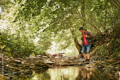 Trekking with backpack concept image. Backpacker in trekking boots crossing mountain river. Man hiking in mountains during summer trip