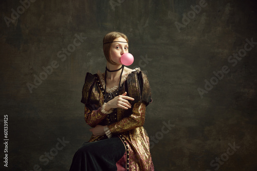 Portrait of pretty young girl, medieval princess in vintage dress posing with bubble gum against dark green background. Concept of history, renaissance art remake, comparison of eras, modernity