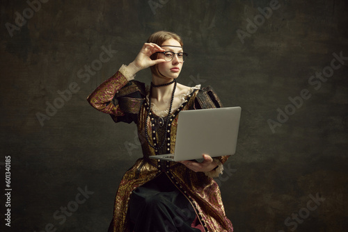 Portrait of young girl, royal person, princess in vintage dress and modern glasses working on laptop against dark green background. Concept of history, renaissance art remake, comparison of eras