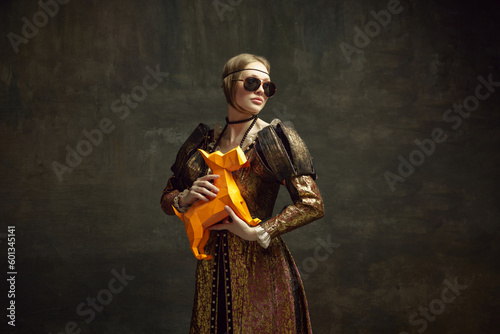 Portrait of young stylish girl in vintage dress and modern sunglasses posing with paper figure of dog against dark green background. Concept of history, renaissance art remake, comparison of eras
