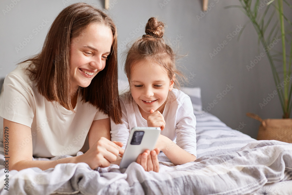Smiling mother and adorable daughter using phone, relaxing on bed at home, little girl child with mum looking at smartphone screen with happy expression. Smiling mother and adorable daughter using phone, relaxing on bed at home, little girl child with mum looking at smartphone screen with happy expression.