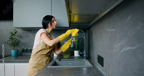 Caucasian young woman in protective yellow gloves washing the kitchen sink and faucet. Housework and housekeeping concept photo