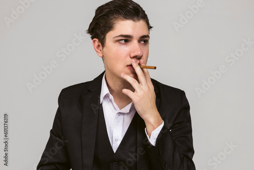 Portrait of a young mafia member with a slicked back hair, dressed in black suit smoking a cigar, throws a menacing gaze. Italian mafia gangster style. isolated on white background.