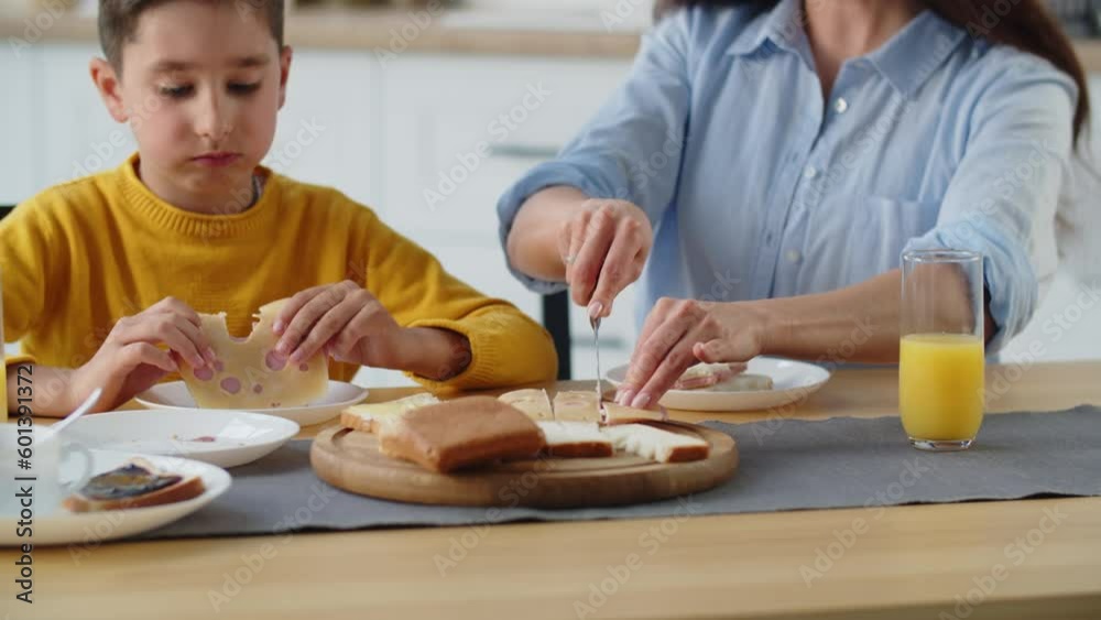 A long-haired brunette woman sits at a table and cuts bread to make sandwiches for her son. A boy in a yellow sweater eats a big cheese sandwich while sitting in the kitchen with his mother.