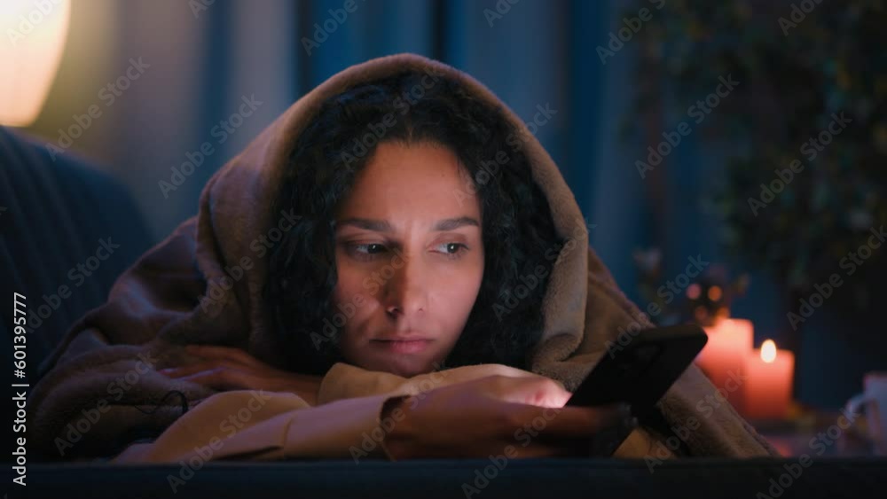 Hispanic sad sleepy girl Latino tired woman upset boring Indian lady bored Arabian female night evening dark home under blanket cover with duvet scrolling mobile phone addict smartphone social media