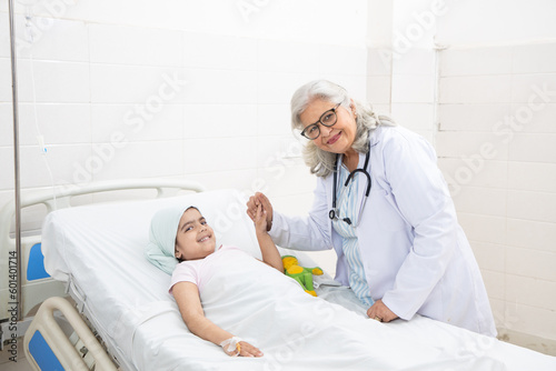 Smiling indian senior woman doctor holding hand of little girl cancer patient lying on hospital bed undergoing course of chemotherapy. Looking at camera.