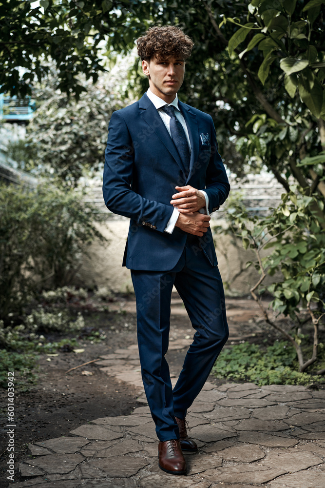 Handsome young man in blue suit posing in green garden Handsome young man in blue suit posing in green garden