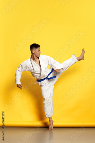 Portrait of professional male taekwondo, karate athlete wearing white kimono doing basic movements over yellow background. Foot kicking