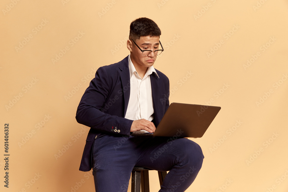Portrait with one handsome businessman, office clerk wearing fashion style costume, suit looking on laptop screen with upset face over beige background Portrait with one handsome businessman, office clerk wearing fashion style costume, suit looking on laptop screen with upset face over beige background