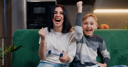 Cute boy with mather sitting on couch and playing video games. Boy and woman uses PlayStation holding console. Games at home together, mom and son. Happy family