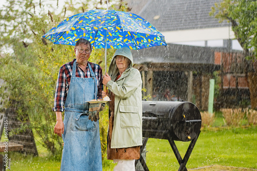 Quadro su tela Frustrated senior couple under an umbrella as rain ruins their outdoor barbecue party