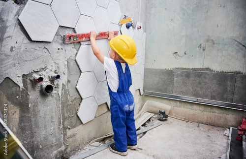 Little boy checking wall surface with spirit level in apartment. Kid in work overalls using level instrument while working on home renovation.