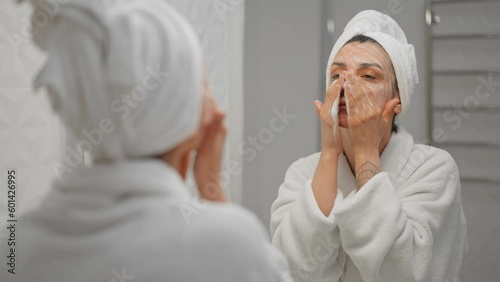Woman using moisturizing foam for face washing and smiling, morning skincare routine. Young female student wearing towel on head looking in mirror in spa salon. Preparation for make up