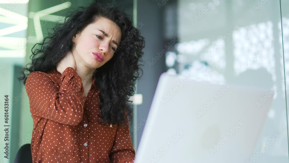 Tired curly brunette woman suffering from neck pain while working on laptop while sitting at desk at workplace in modern office. Sad female employee massages and rubs sore muscles, stretches