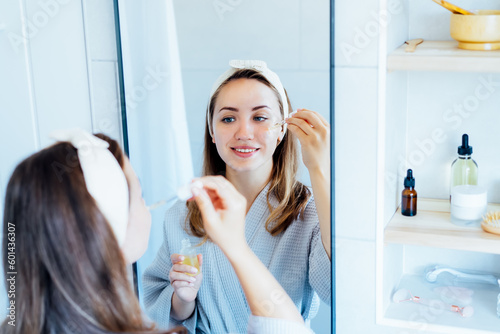 Young woman in bathrobe looking in the mirror, holding dropper with hyaluronic acid serum close to face and smiling. Skin hydrating. Cosmetic spa procedures. Beauty self-care at home. Selective focus.