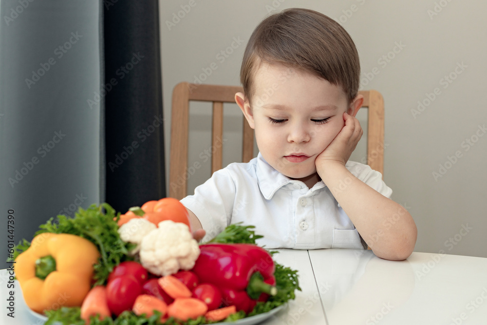 kid doesn't like healthy food concept, sad, unhappy boy next to a dish of fresh vegetables kid doesn't like healthy food concept, sad, unhappy boy next to a dish of fresh vegetables