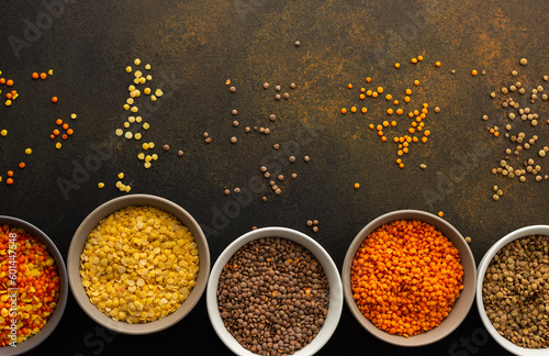 Multi-colored lentils in bowls on a brown background, scattered yellow and brown, green and orange lentils, healthy legumes, top view