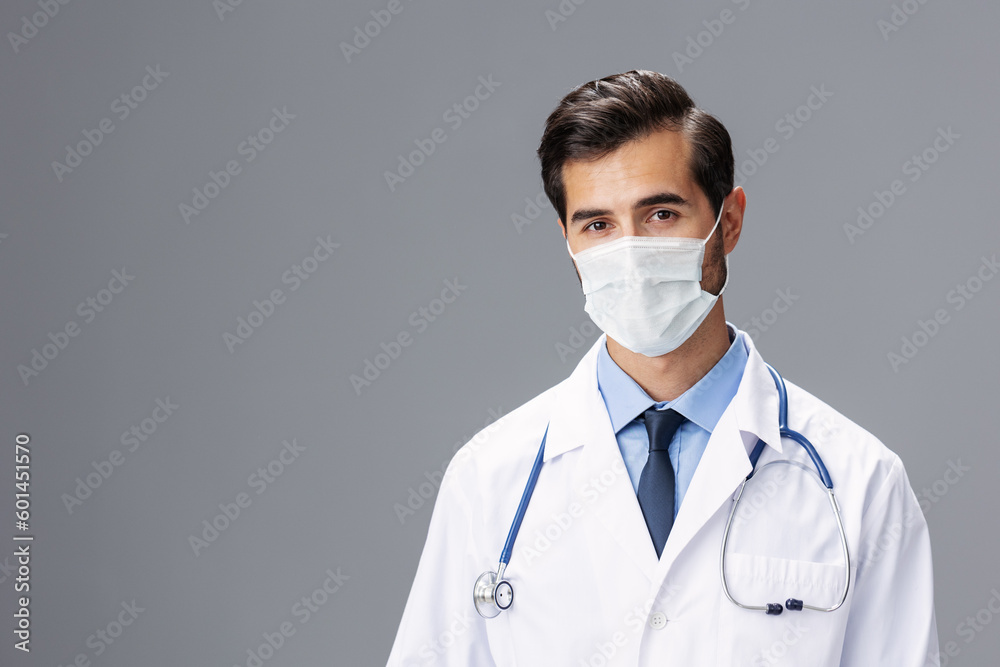 A male doctor in a white coat and medical mask and sterile gloves looks at the camera on a gray isolated background, copy space, space for text