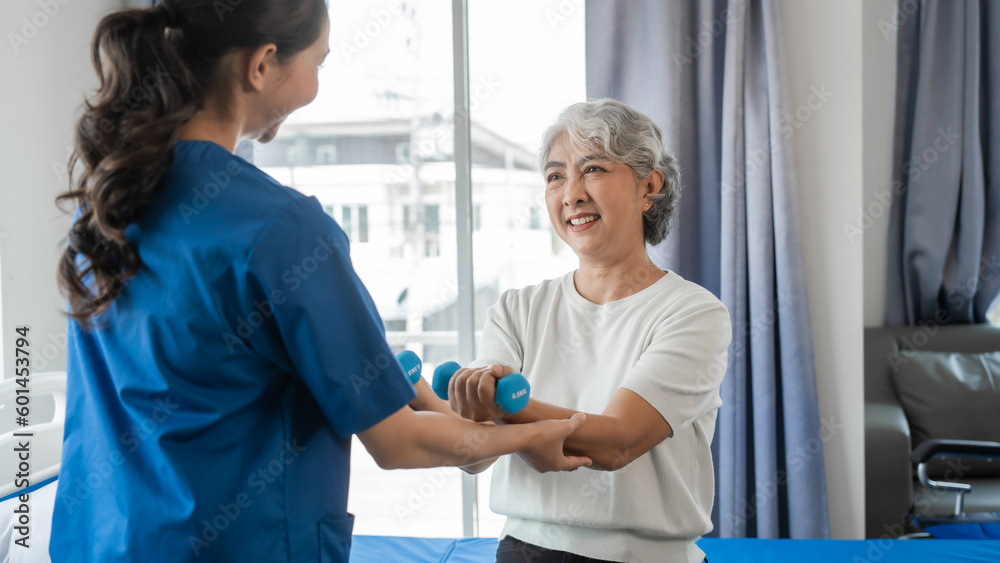 Obraz premium Young physiotherapist helping senior mature asian woman grey hair work out with dumbbells, to recover from injury at health centre in physical therapy session.