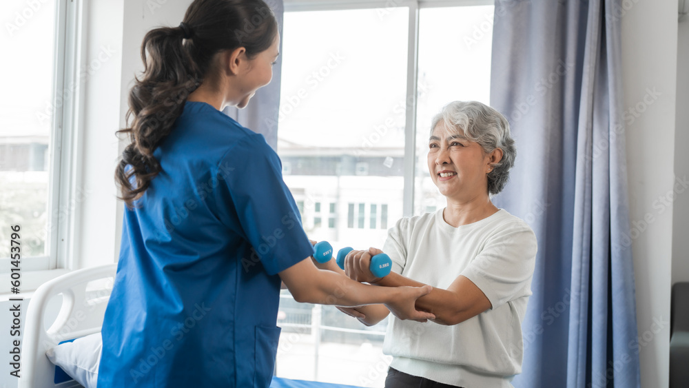 Obraz premium Young physiotherapist helping senior mature asian woman grey hair work out with dumbbells, to recover from injury at health centre in physical therapy session.