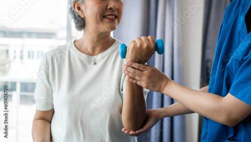 Fotografie, Obraz Young physiotherapist helping senior mature asian woman grey hair work out with dumbbells, to recover from injury at health centre in physical therapy session