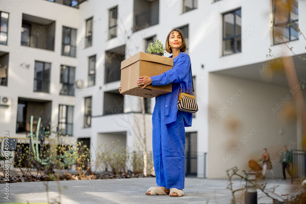 Young woman carrying cardboard box and flowerpot at inner yard of apartment building, relocating to a new home