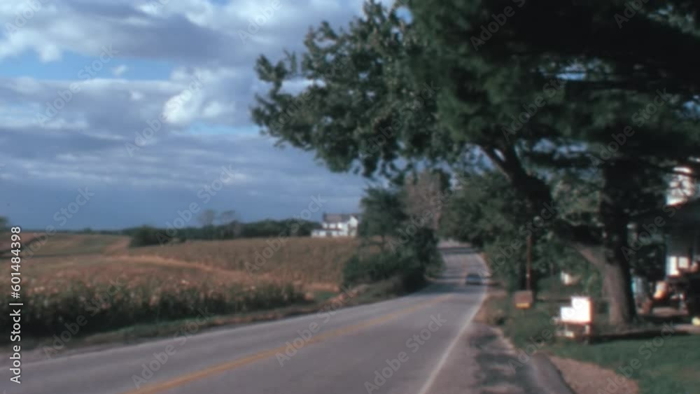 A quiet street lined with lush green trees runs alongside a vast wheat field and a dramatic cloudy sky in the background. Vintage footage of Baltimore, Maryland in 1960. 8mm video effect. A quiet street lined with lush green trees runs alongside a vast wheat field and a dramatic cloudy sky in the background. Vintage footage of Baltimore, Maryland in 1960. 8mm video effect.