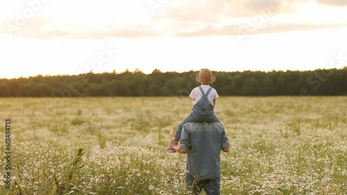 A young dad carries a little daughter in a straw hat in her arms. Walking through the chamomile field at sunset. The concept of life outside the city and walks outside the city on the weekend.