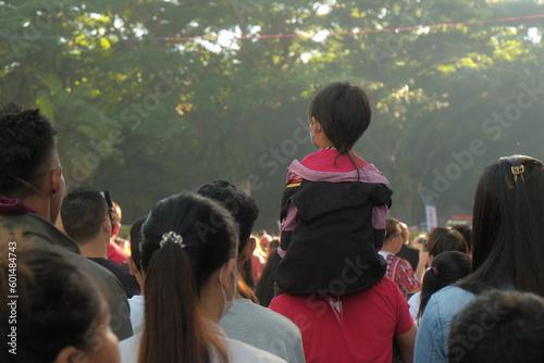 Malaybalay, Philippines - father carries his son, a young boy with a rattail, on his shoulders among a busy crowd at the Kaamulan Festival in Bukidnon. Post-pandemic outdoor event. People from behind. photo