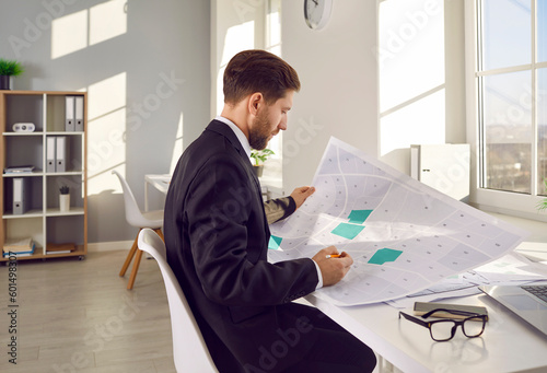 Side view portrait of a professional cartographer working with printed cadastral map at table on his workplace. Young man analyzing cadastral map and searching for a building plot. photo