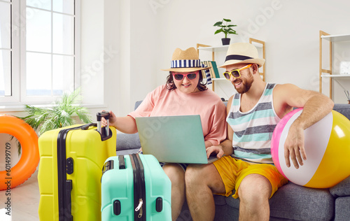 Happy family couple in summer hats and sunglasses sitting on sofa with beach ball and travel bags, using laptop computer, and booking tickets and holiday accommodation online