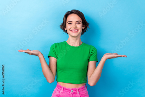 Photo of cheerful positive woman wear green t-shirt smiling comparing arms empty space isolated blue color background