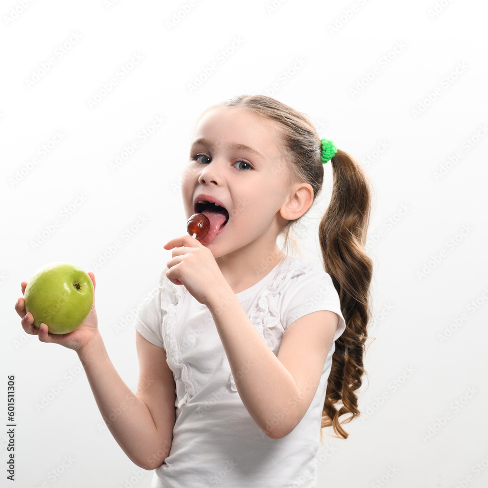 A little girl licks a lollipop and holds an apple in her hand. A little girl licks a lollipop and holds an apple in her hand.