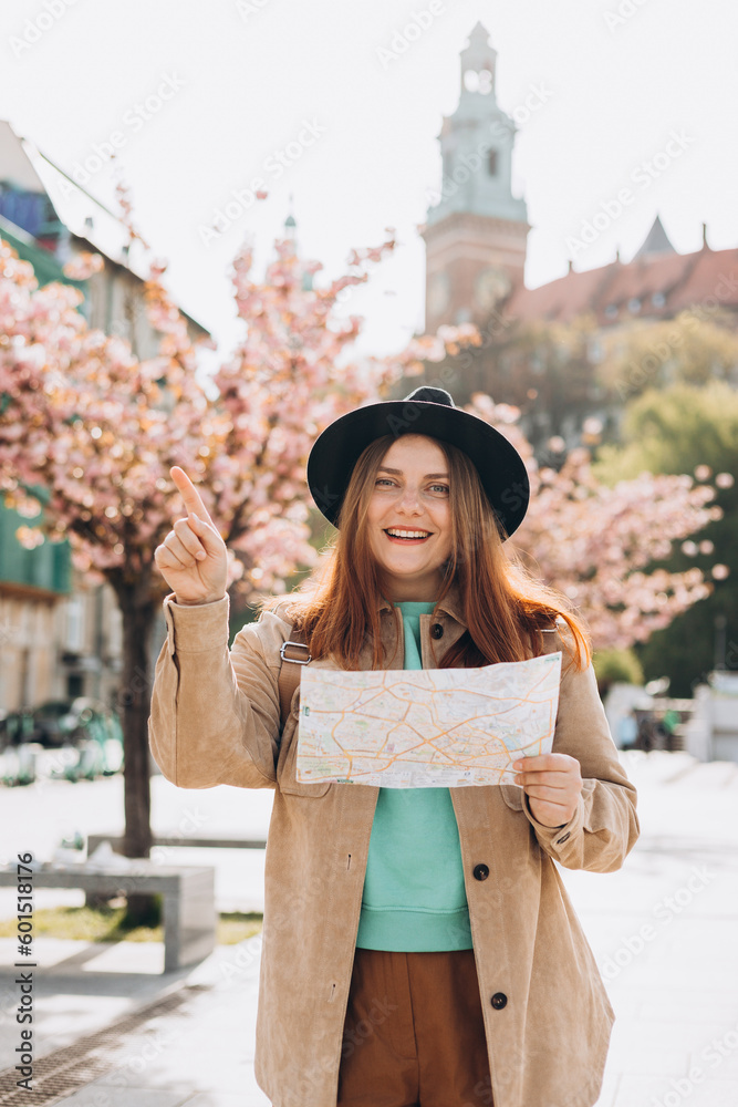 Attractive female tourist is exploring new city. Happy Redhead woman pointing finger and holding a paper map in Krakow. Traveling Europe in spring. Active lifestyle